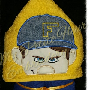 May include: Yellow hooded towel with a blue and white embroidered face wearing a blue denim baseball cap with the letter "F" on it. The towel has a yellow ribbon tied around it.
