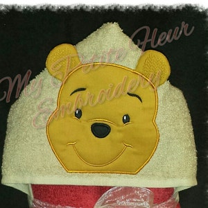 Puede incluir: Una toalla con capucha roja con un diseño bordado de Winnie the Pooh en la capucha. La toalla está atada con una cinta blanca y tiene un tarro de miel bordado en la parte delantera con la palabra "Hunny" en él.