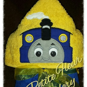 May include: A yellow hooded towel with a blue and gray embroidered train engine on the hood. The towel is tied with a yellow ribbon and the words "My Petite Fleur Embroidery" are embroidered on the towel.