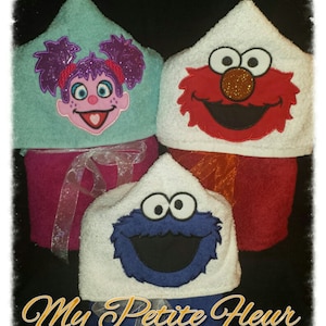May include: Three hooded towels with embroidered characters from Sesame Street. The towels are white, turquoise, and red. The characters are Elmo, Cookie Monster, and Abby Cadabby. The towels are stacked on top of each other. The text "My Petite Fleur Embroidery" is at the bottom of the image.