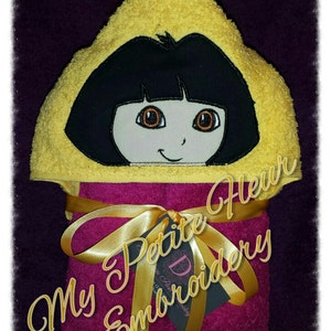 May include: A pink and yellow hooded towel with an embroidered image of Dora the Explorer. The towel is tied with a gold ribbon and has a tag that says "My Petite Fleur Embroidery".