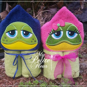 May include: Two hooded towels with frog faces embroidered on them. The towels are rolled up and tied with ribbons. The frog on the left towel has blue eyes and a blue ribbon. The frog on the right towel has pink eyes and a pink ribbon.