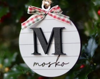 May include: White round Christmas ornament with a black letter M and the name "mosko" written in black cursive script. The ornament is decorated with a red and white gingham ribbon.