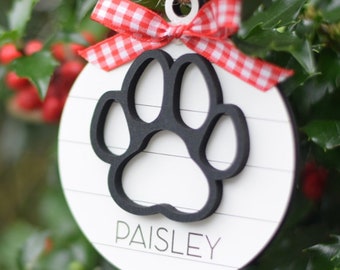 May include: White wooden Christmas ornament with a black paw print and the name "PAISLEY" written in black. The ornament is decorated with a red and white gingham bow.