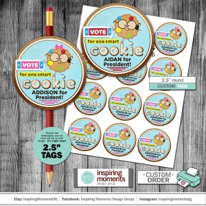 May include: Round 6.35 cm custom tags featuring a cartoon cookie character with glasses and a bow. The tags read "VOTE for one smart cookie, Addison for President!" and "Aidan for President!" A pencil and printed tags are visible.