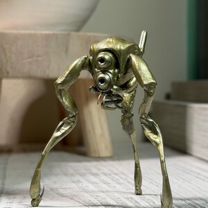 Puede incluir: Escultura de latón de una criatura mecánica con tres patas, similar a un insecto. Presenta dos grandes elementos circulares en forma de ojos y una pequeña antena. Las patas son largas y delgadas, con una ligera curvatura en las rodillas.
