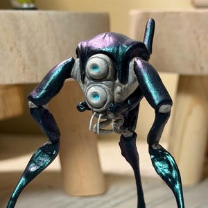 Puede incluir: Una criatura robótica metálica de color verde azulado y púrpura con grandes ojos redondos y azules. La figura tiene múltiples extremidades articuladas y un diseño detallado similar a un insecto. La criatura se encuentra sobre una superficie texturizada.