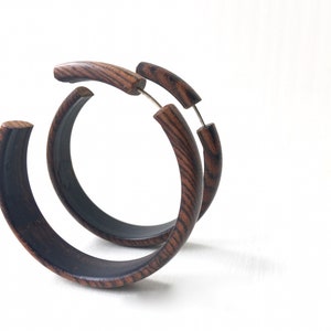 Könnte beinhalten: Ein Paar große, dunkelbraune Holz-Creolen. Die Ohrringe sind aus dunklem Holz mit natürlicher Maserung. Die Creolen sind dick und haben eine leicht abgerundete Form.