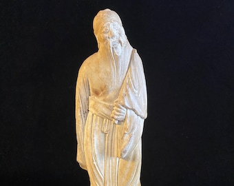 Op de afbeelding: Beige beeld van een oude man met een lange baard en traditionele kleding, staand op een kleine, donkerbruine houten standaard. Het beeld heeft gedetailleerde gelaatstrekken en staat tegen een zwarte achtergrond.
