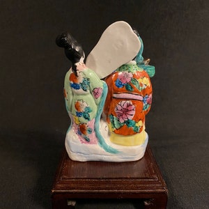 May include: A ceramic figurine depicting two figures viewed from the back, featuring a light green robe with floral patterns and an orange robe with pink and yellow floral designs. The figures stand on a white base atop a dark brown wooden stand.