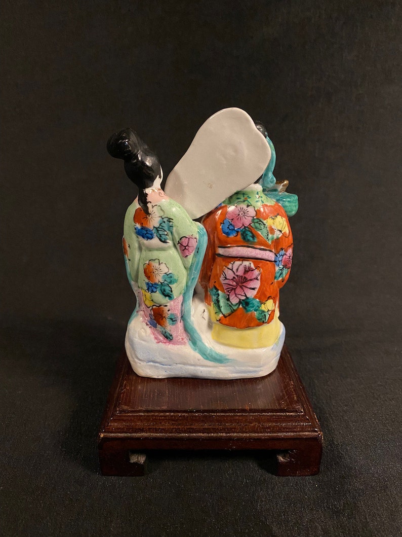 May include: A ceramic figurine depicting two figures viewed from the back, featuring a light green robe with floral patterns and an orange robe with pink and yellow floral designs. The figures stand on a white base atop a dark brown wooden stand.