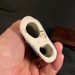 May include: A hollow ceramic figurine base featuring two distinct oval-shaped openings. The underside is off-white with visible brown discoloration and signs of wear around the edges, showing the interior structure of the object.
