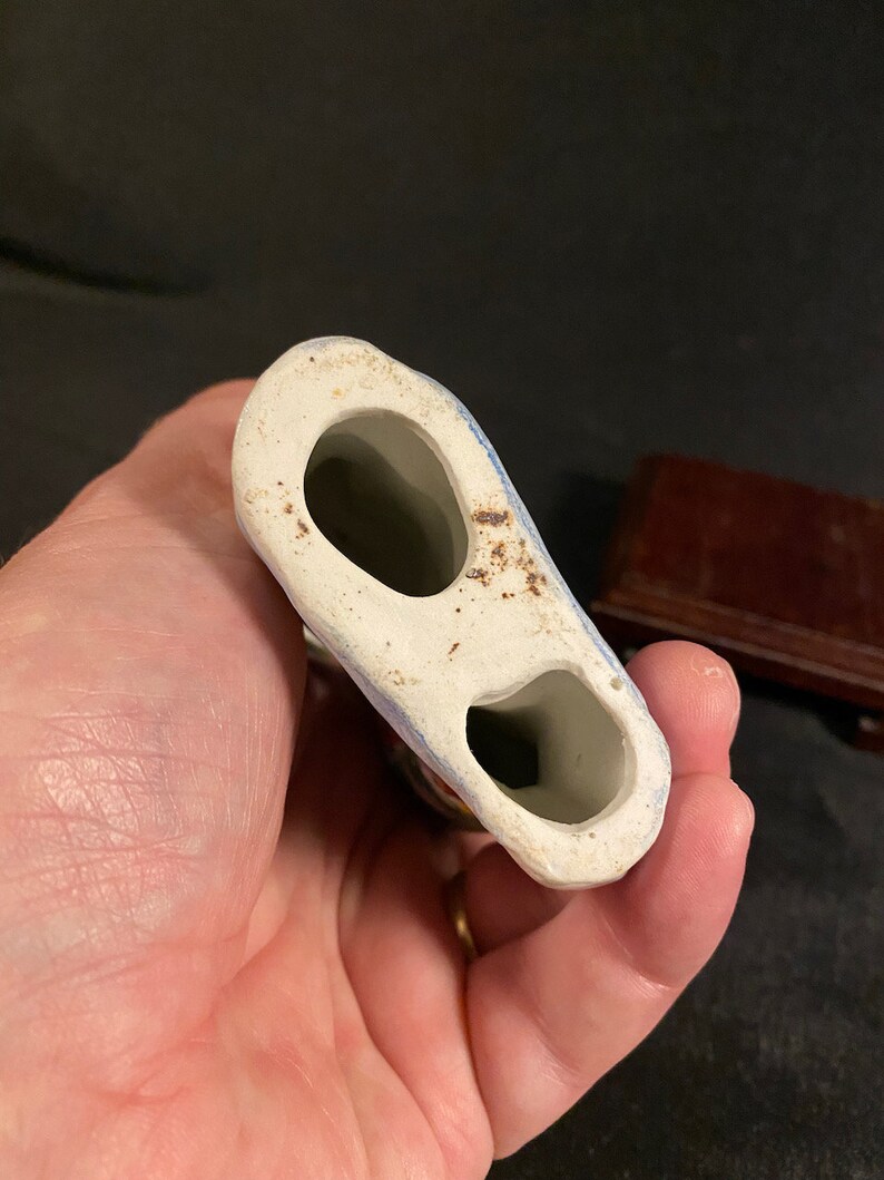 May include: A hollow ceramic figurine base featuring two distinct oval-shaped openings. The underside is off-white with visible brown discoloration and signs of wear around the edges, showing the interior structure of the object.