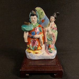May include: A porcelain figurine featuring two figures in traditional attire standing on a white base. One figure wears an orange floral robe and holds a green tray, while the other wears a green robe and holds a blue and white hand fan.
