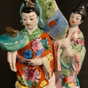 May include: A porcelain figurine featuring two figures in traditional attire. One figure wears an orange robe with floral patterns and holds a teal circular object, while the other wears a light green robe and holds a large, fan-shaped accessory.