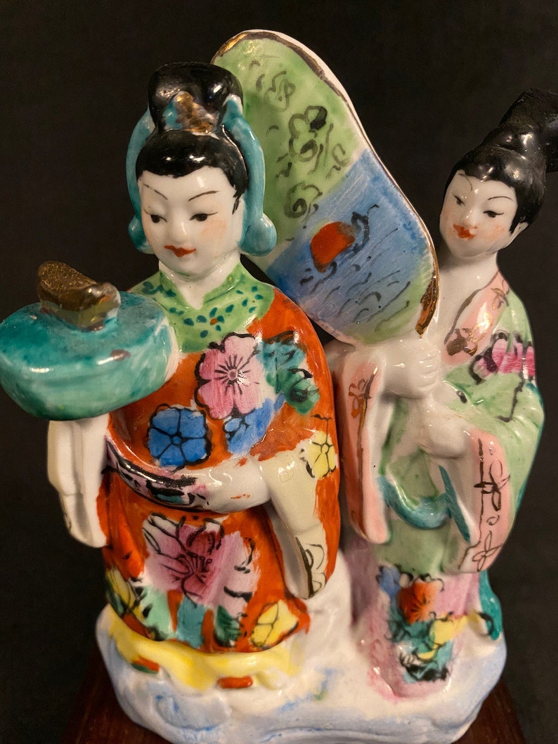 May include: A porcelain figurine featuring two figures in traditional attire. One figure wears an orange robe with floral patterns and holds a teal circular object, while the other wears a light green robe and holds a large, fan-shaped accessory.