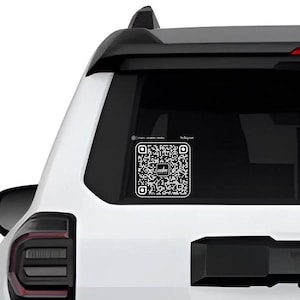 May include: A white car with a black QR code sticker on the back window. The QR code has the text "adm" in the center.