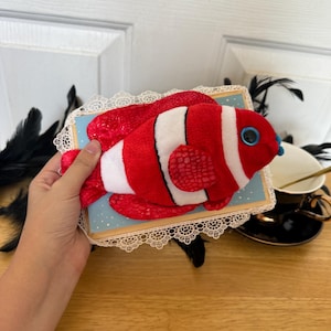 May include: A plush clownfish toy with red, white, and black details. The fish is mounted on a light blue rectangular base with a white lace trim. The toy has blue eyes and is held by a person.