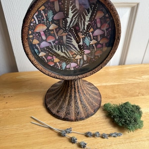 May include: A circular, bronze-toned metal display stand with a textured, fluted base and frame. The center features a glass-covered panel displaying a black and white butterfly graphic layered over a dark background with colorful mushrooms.