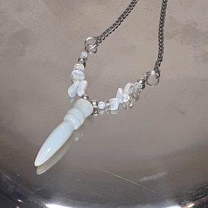 Può includere: Una delicata collana con una catena d'argento e un ciondolo azzurro. Il ciondolo ha un design scolpito. La collana presenta anche piccole perle bianche irregolari e dettagli argentati. Un accessorio elegante.