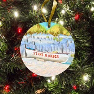 May include: A round, flat Christmas ornament featuring a painted scene of a white boat with the text STONE HARBOR in red letters. The boat rests on a sandy beach in front of a wooden fence and tall yellow beach grass under a blue sky.
