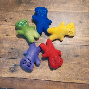 May include: Five colorful felt figures in blue, green, yellow, purple, and red, each with embroidered facial features. The figures are arranged on a wooden surface, creating a playful and vibrant composition. These plush toys are ideal for children.