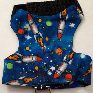 May include: A blue pet harness with a space-themed pattern featuring rockets, planets, and stars. The harness has a black strap and a metal D-ring for leash attachment. The rockets have red and silver details.