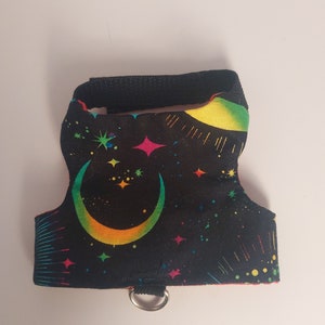Puede incluir: Un pequeño arnés para mascotas con un fondo negro y un diseño celestial colorido. El arnés presenta una luna creciente, estrellas y acentos de arcoíris. Tiene una correa negra y un anillo metálico para sujetar la correa.