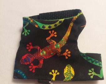 May include: Black fabric pet harness featuring a colorful lizard design. The lizard is depicted in red, orange, green, and yellow geometric patterns. The harness has a black strap and a small metal ring.