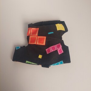 Puede incluir: Un arnés negro para mascotas con un patrón geométrico colorido. El patrón incluye cuadrados en rojo, naranja, rosa, amarillo y azul. El arnés tiene una correa negra y una hebilla pequeña.