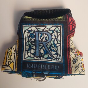Può includere: Un'imbracatura per animali domestici con un design a vetrata, con la scritta "RAVENCLAW" e una grande "R" blu. Ha una cinghia nera e un anello di metallo per attaccare il guinzaglio. Il tessuto è a fantasia colorata.