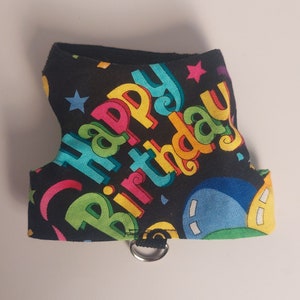 Puede incluir: Un arnés negro para mascotas con las palabras "Happy Birthday" en letras coloridas. El arnés tiene un anillo en D para sujetar la correa y está decorado con estrellas y globos.