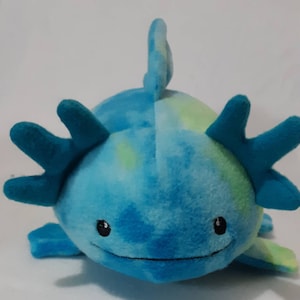 May include: A plush axolotl toy in shades of blue and green. The stuffed animal has a rounded body, a fin on its back, and six frilly gills. It has small, black, round eyes and a smiling mouth.