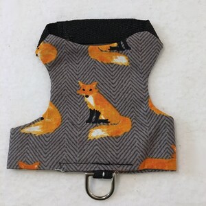 May include: A gray and orange fox-themed pet harness. The harness features a repeating pattern of orange foxes on a gray herringbone background. It has a black strap and a metal D-ring for leash attachment.