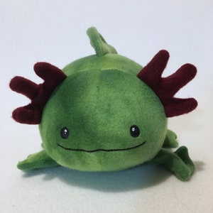 May include: A plush toy shaped like an axolotl with a round green body, dark red feathery external gills on each side of its head, and a small dorsal fin on top. The face features two small black embroidered eyes and a simple horizontal line for a mouth.