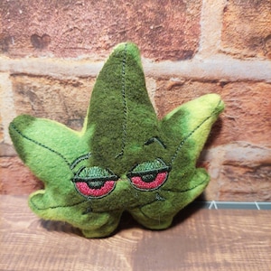 May include: A plush, green leaf with embroidered red eyes and a sleepy expression. The leaf has a stitched outline and a yellow-green gradient. The item is likely a novelty item or a decorative piece.