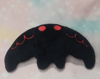 Puede incluir: Un peluche negro con forma de polilla con detalles bordados en rojo. La polilla tiene dos ojos circulares rojos y líneas decorativas rojas en las alas. El juguete está hecho de material suave.