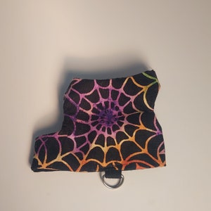 Puede incluir: Un pequeño arnés negro para mascotas con un diseño de telaraña colorido en tonos morado, naranja y amarillo. El arnés tiene un anillo en D para la fijación de la correa.