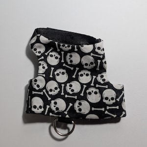 May include: A black and white pet harness with a repeating pattern of white skulls and crossbones. The harness has a black strap and a silver metal ring for leash attachment. This pet accessory is designed for small animals.