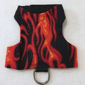 May include: A small pet harness featuring a flame design in red and yellow against a black backdrop. It has a black strap and a silver metal D-ring for leash attachment. The harness is designed for small pets.