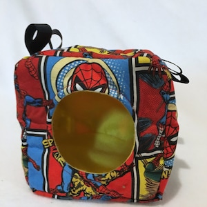 May include: A square fabric toy with a Spiderman comic book design. It has a large, circular opening and a black loop at the top. The fabric is red, blue, and yellow, with images of Spiderman.