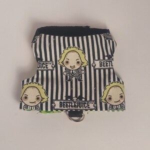 May include: Black and white striped pet harness featuring cartoon images of Beetlejuice. The harness has a black trim and a metal D-ring for leash attachment. The text "BEETLEJUICE" is printed on the harness.