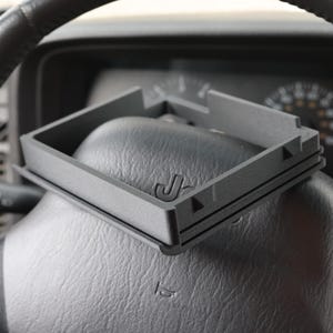 May include: A dark gray, square-shaped car dashboard accessory with the letter "J" embossed on its surface. The accessory is resting on a black steering wheel, with the car's dashboard and gauges visible in the background.