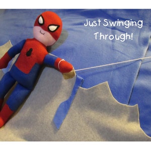 May include: A red and blue felt Spiderman doll is swinging on a white string. The doll has a black spider logo on its chest. The background is a blue and gray felt fabric. The text "Just Swinging Through!" is written in white on the blue fabric.