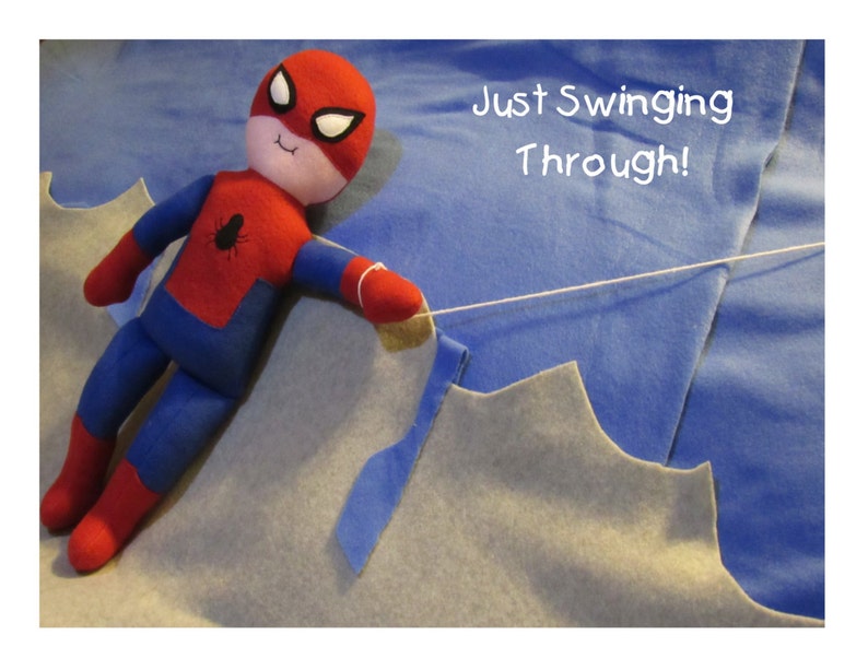 May include: A red and blue felt Spiderman doll is swinging on a white string. The doll has a black spider logo on its chest. The background is a blue and gray felt fabric. The text "Just Swinging Through!" is written in white on the blue fabric.