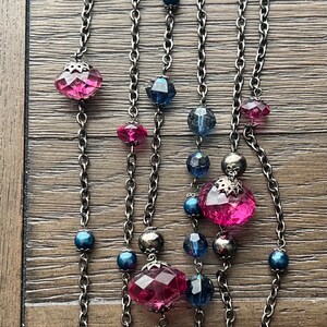 May include: A silver chain necklace with pink and blue faceted glass beads. The necklace has a layered look with multiple strands of chain.