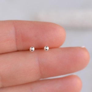 May include: A pair of small, silver ball stud earrings. The earrings are shown on a person's finger.