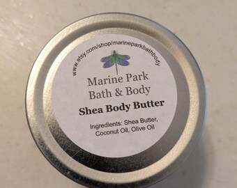 Puede incluir: Una lata de plata con una etiqueta blanca que dice "Marine Park Bath & Body Shea Body Butter" con una ilustración de libélula y los ingredientes que se enumeran a continuación: Manteca de karité, Aceite de coco, Aceite de oliva.