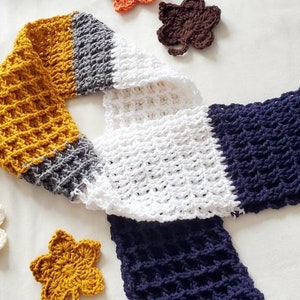 May include: A crocheted scarf in four colors: mustard yellow, gray, white, and navy blue. The scarf is made with a waffle stitch pattern.
