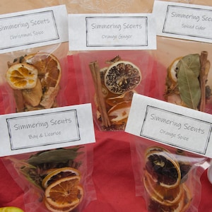 May include: Five clear plastic bags with labels that read "Simmering Scents" and different scents, such as "Christmas Spice", "Orange Ginger", "Spiced Cider", "Bay & Licorice", and "Orange Spice". Each bag contains a variety of dried fruits, spices, and herbs.